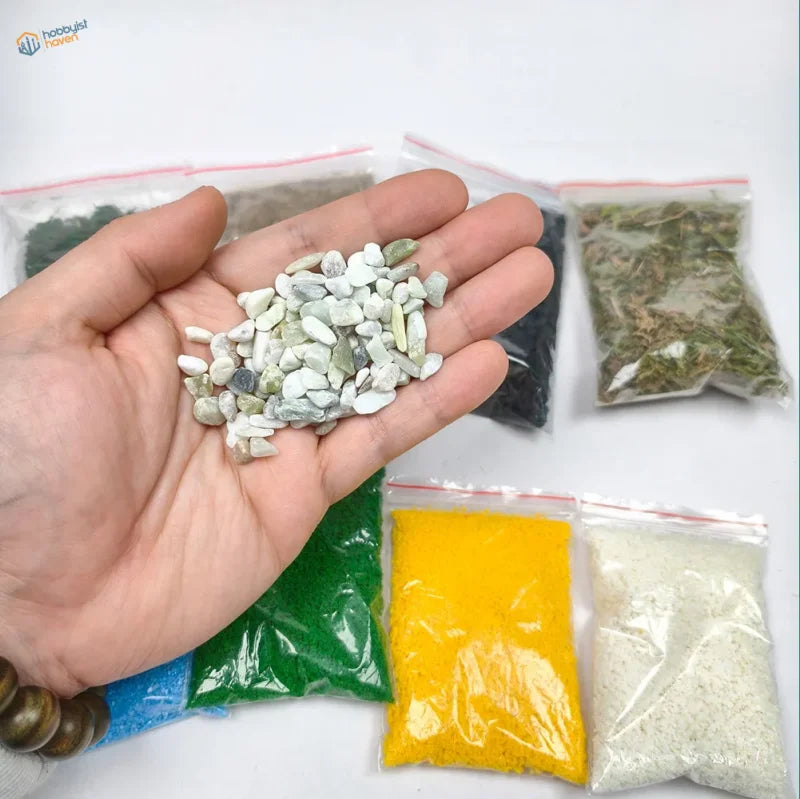 A hand holds smooth light green and white pebbles from a 9pcs diorama scenery material set for scenery starter pack