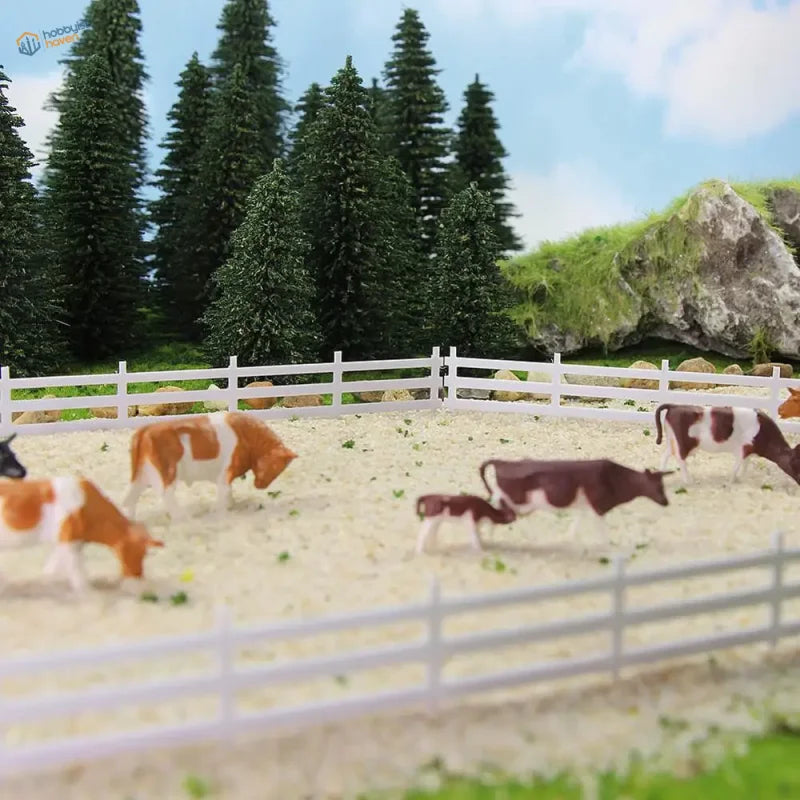 Brown and white plastic cow figurines in a miniature fenced pasture with n scale white lattice-top picket fence panels