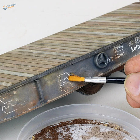 Double-headed weathering brush applying paint to miniature train car rust