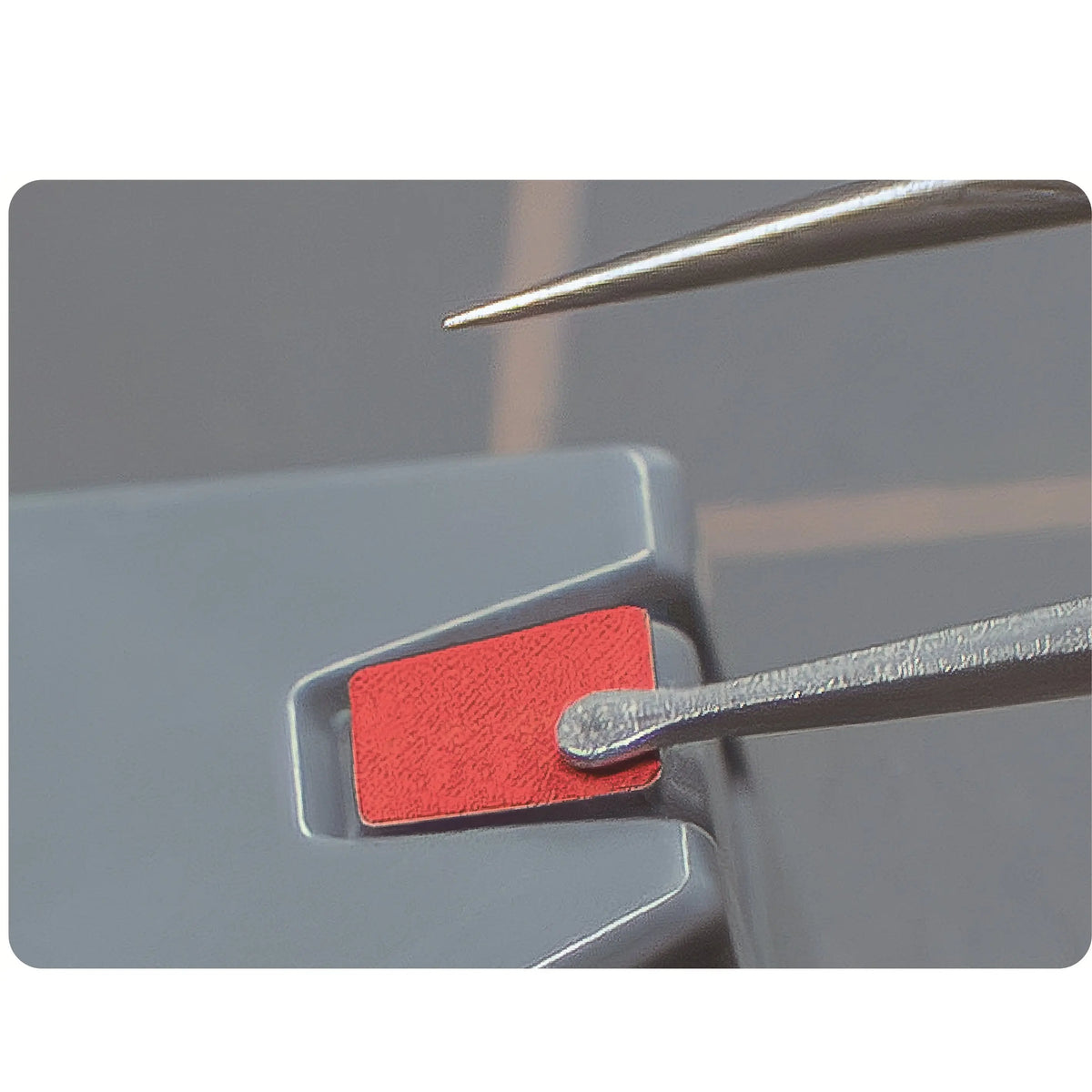 Decal tweezers with a long pointed tip hovering over a decal on a grey model part