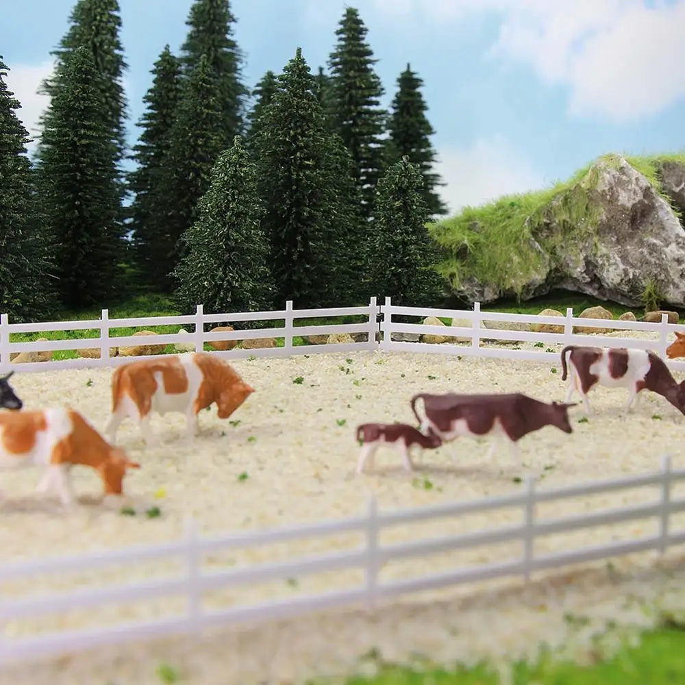 Brown and white plastic cow figurines in a miniature fenced pasture with n scale white lattice-top picket fence panels