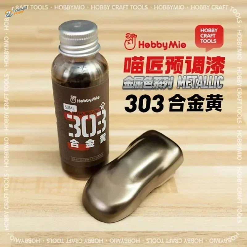 A bottle of Hobby Mio 50ml oil-based metallic paint in 303 gold with a silver cap next to a golden swatch for scale modeling tools