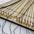 Beige wooden rope ladder weaving jig with black cord threaded through gold pegs and labeled measurement markings